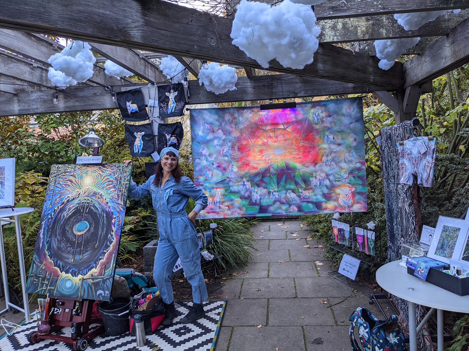 Sari Luna is a live painter and vendor, she is pictured standing with a painting she was working on in front of her booth which features an art installation and her other works.